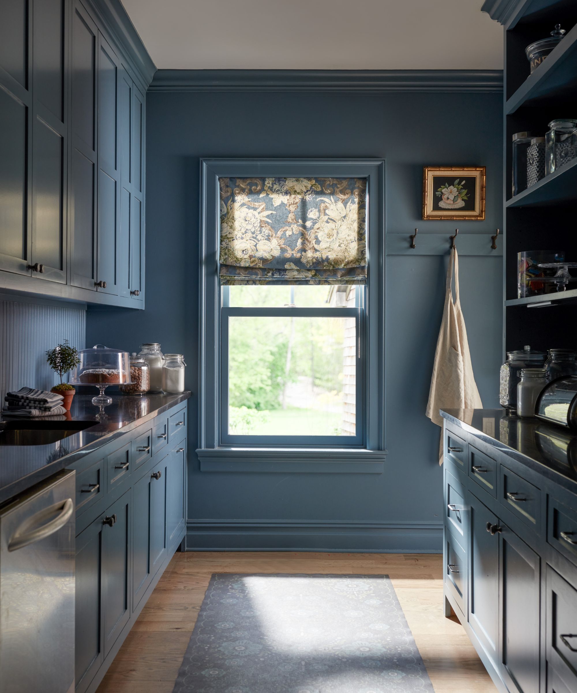 A scullery with dark blue walls, trim, and cabinets, wood flooring with a blue runner rug, a large window with a patterned Roman blind, and coat hooks.