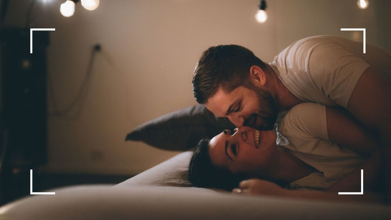 Man lying on top of woman in bed, both laughing and smiling underneath fairy lights, to illustrate the broken eagle sex position