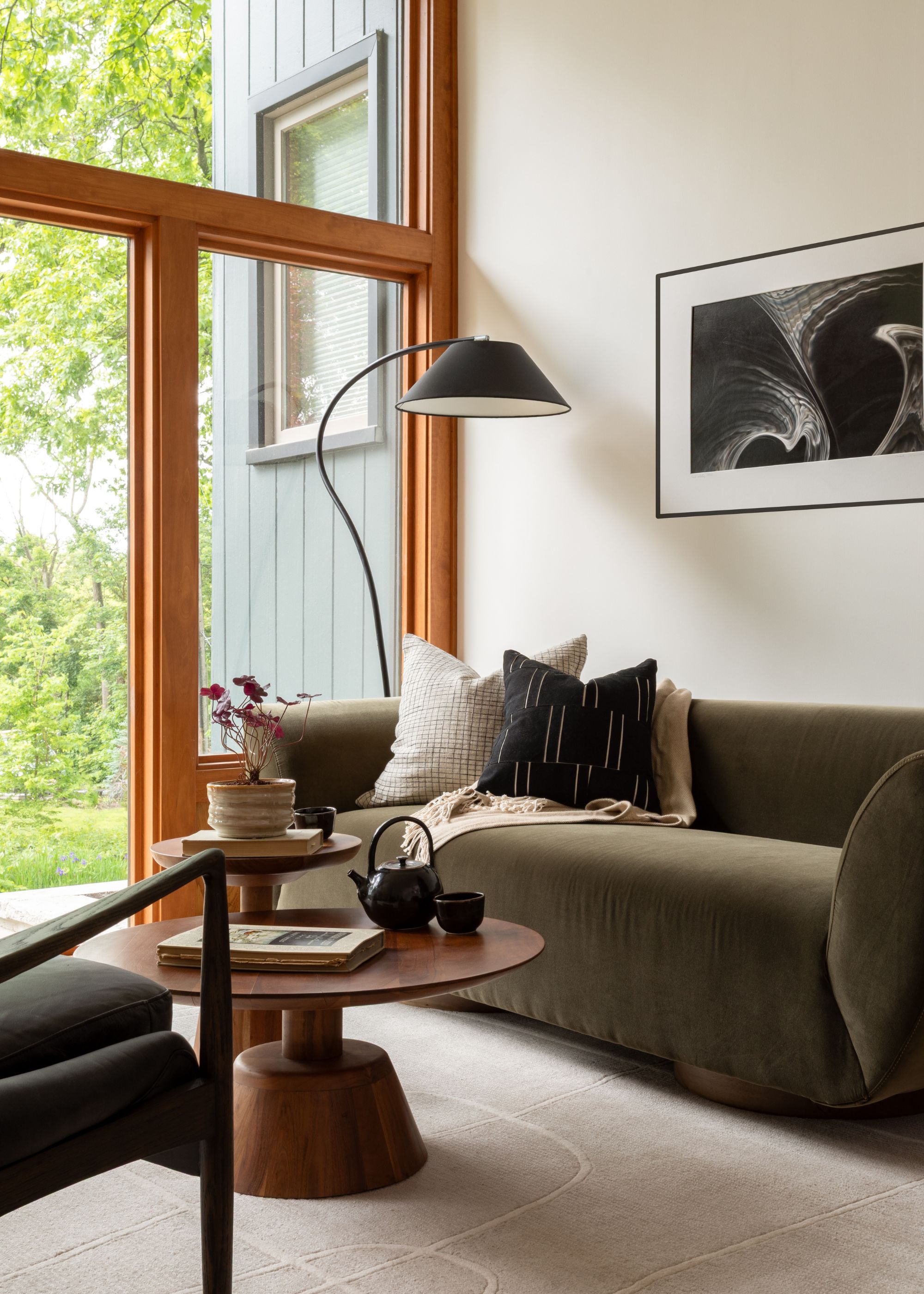 A modern living room with a cream wall with a dark olive green velvet sofa, a wooden coffee table, and a light beige rug. Glass doors leading into a garden and lush green trees in the background.