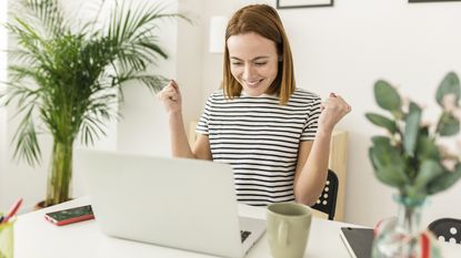 A young woman celebrates as she looks at her open laptop on her desk at home.