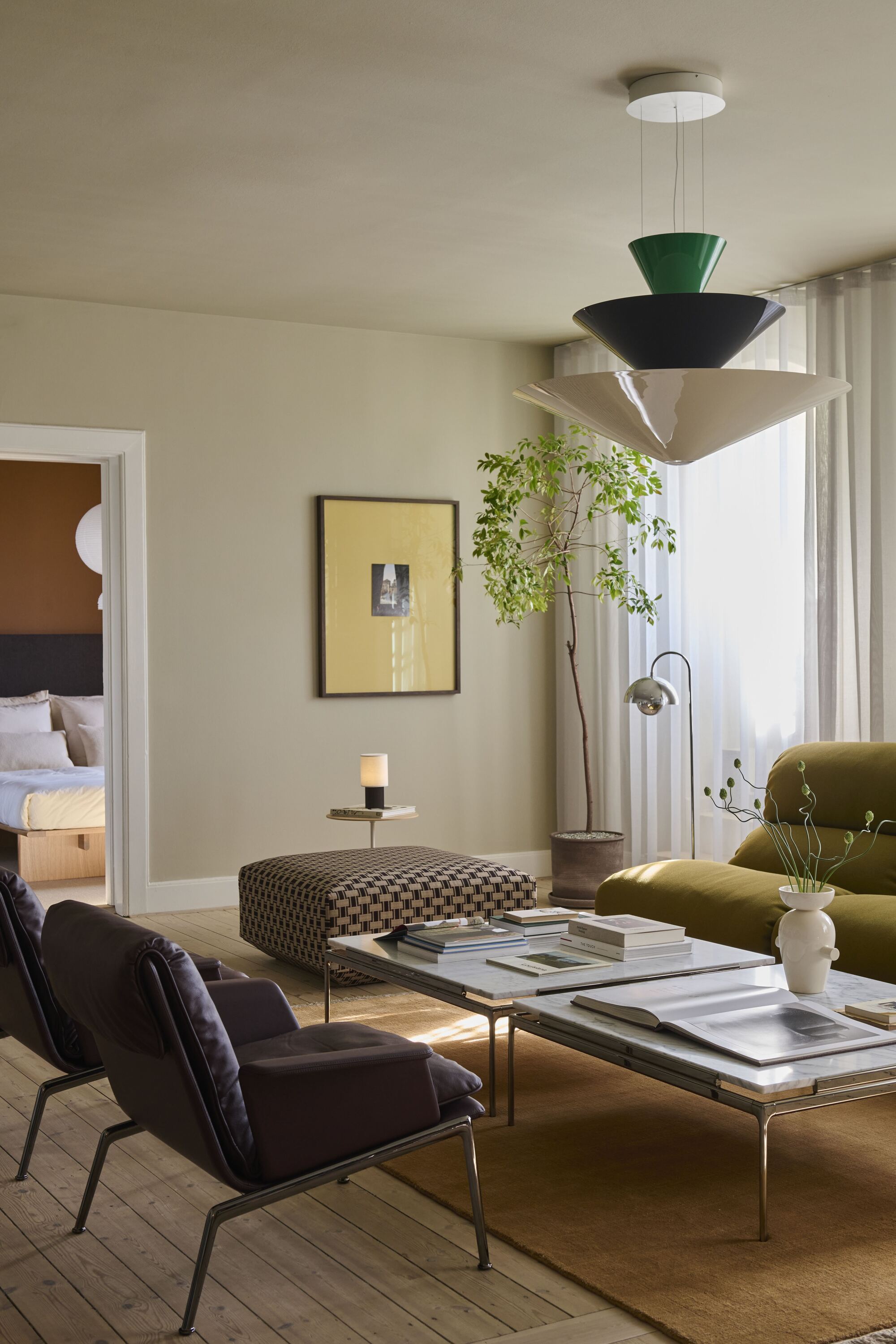 living room with layered lighting, pendant light, floor lamp, and table lamp, two coffee tables on an orange rug, with two armchairs opposite a green sofa, and an ottoman with a side table next to it. in the corner of the room is a potted tree with sheer curtains behind it and an artwork hanging on the wall next to it