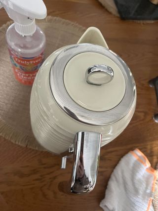 A cream kettle with stainless steel trims on a wooden table next to a cleaning bottle and some cloths