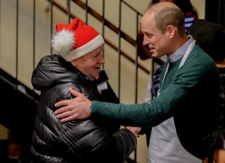 Prince William wearing a green sweater and a white apron embracing a man wearing a santa hat and black coat