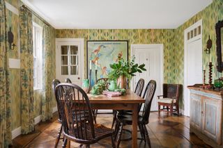 A New England-style decorated living room features warm, pastel-shaded wallpaper, a central dining table in wood, and multiple, aquamarine ceramic vases as well as teapots.