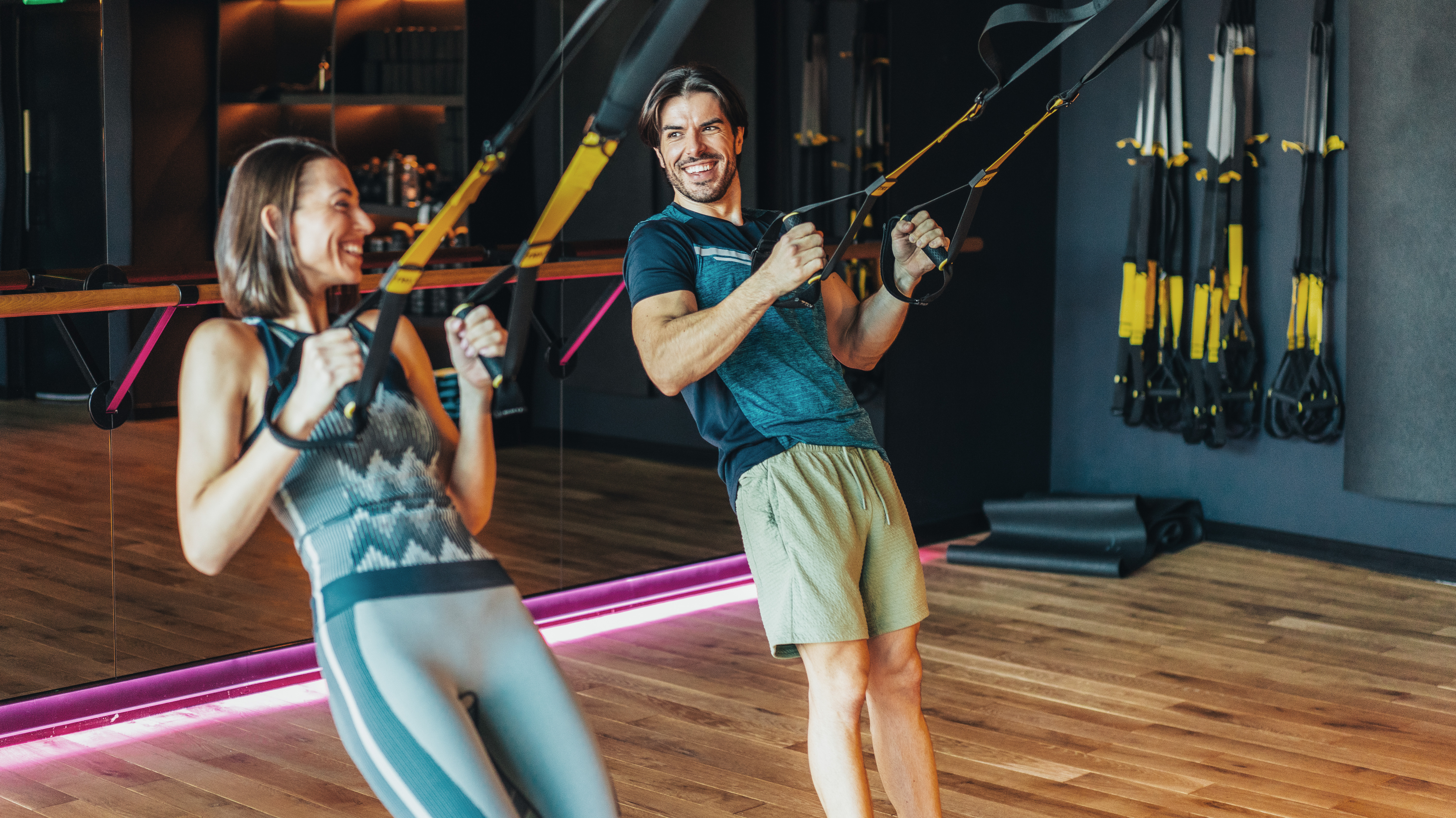 A man and woman in a gym use suspension trainers to perform inverted rows. They are holding onto the straps of the suspension trainer and pulling themselves upward, their elbows bent, both smiling as they exercise.