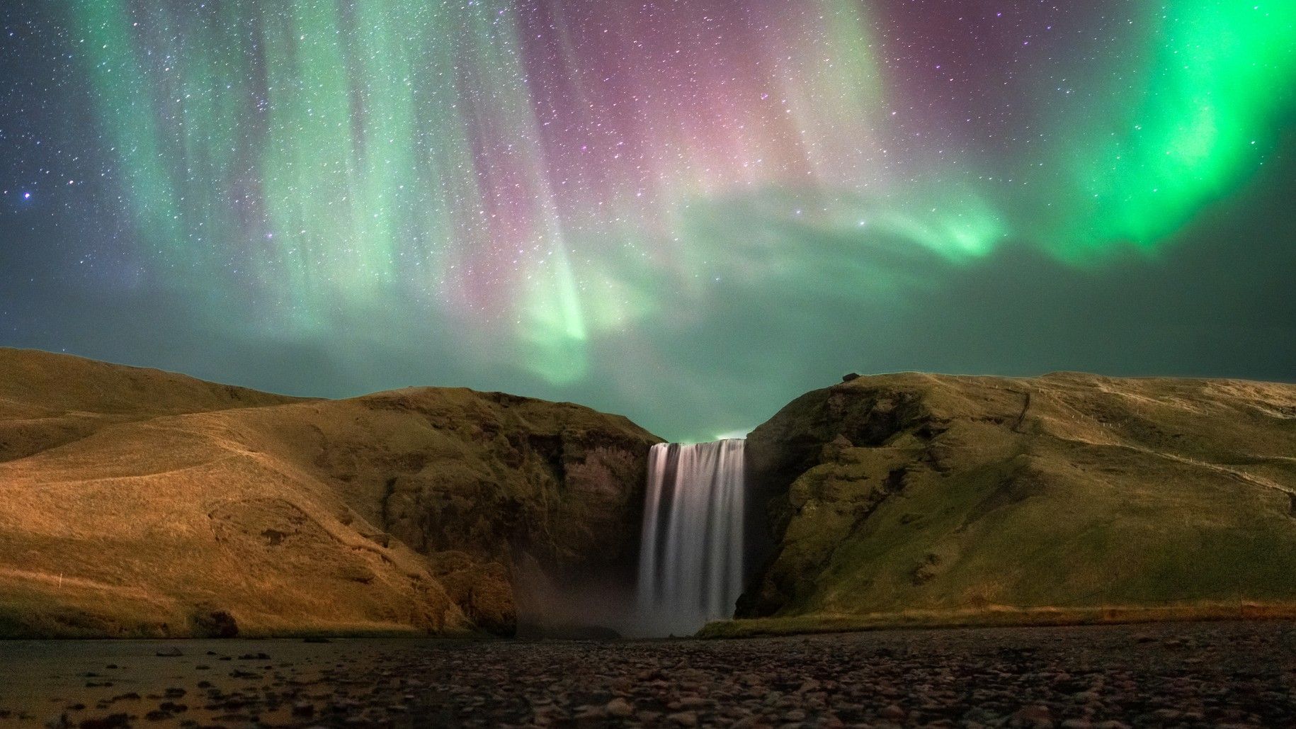 northern lights dance above a large waterfall in Iceland.