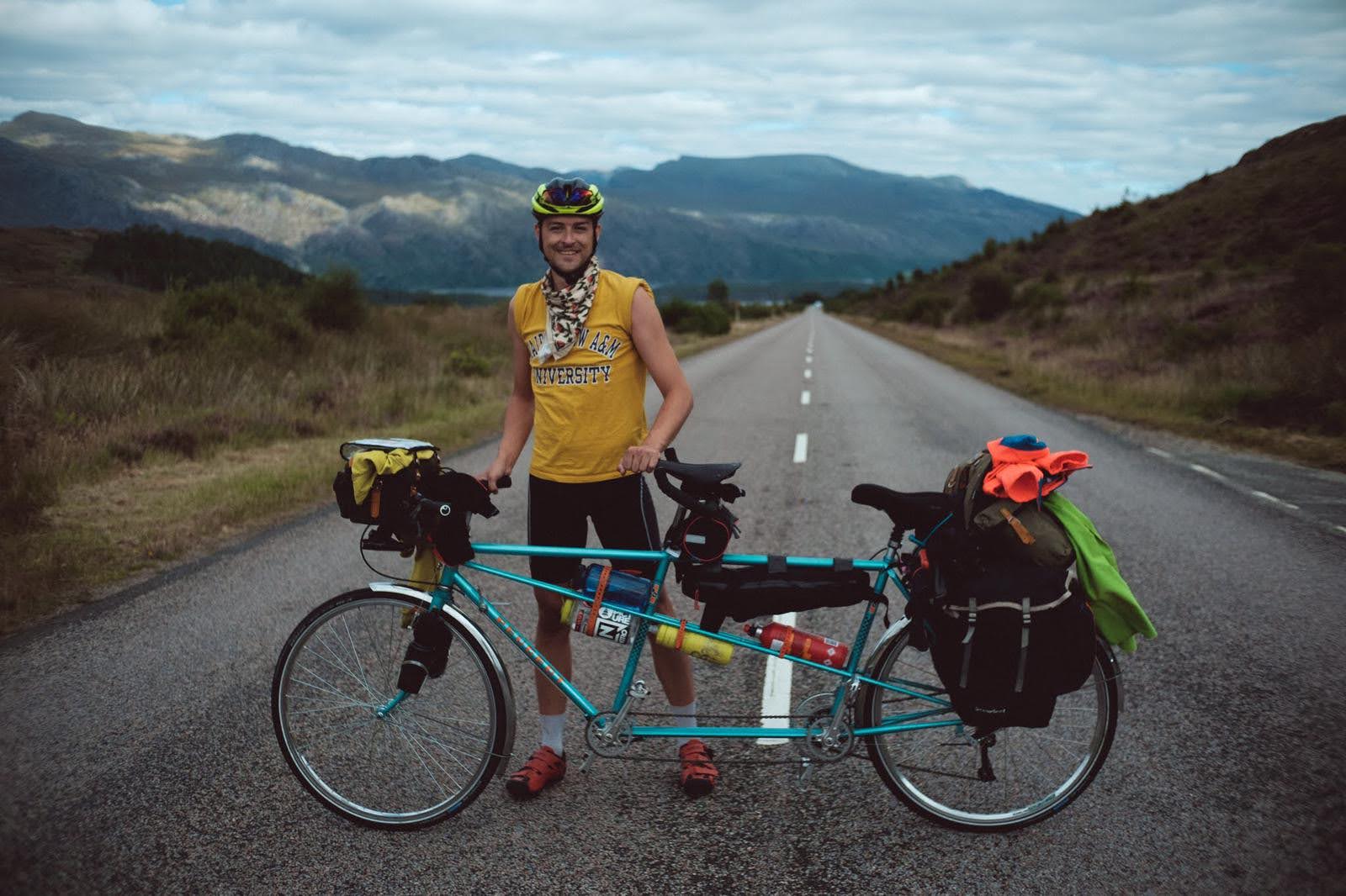 A white man in a yellow t shirt stands behind a heavily laden tandem at the top of a large hill