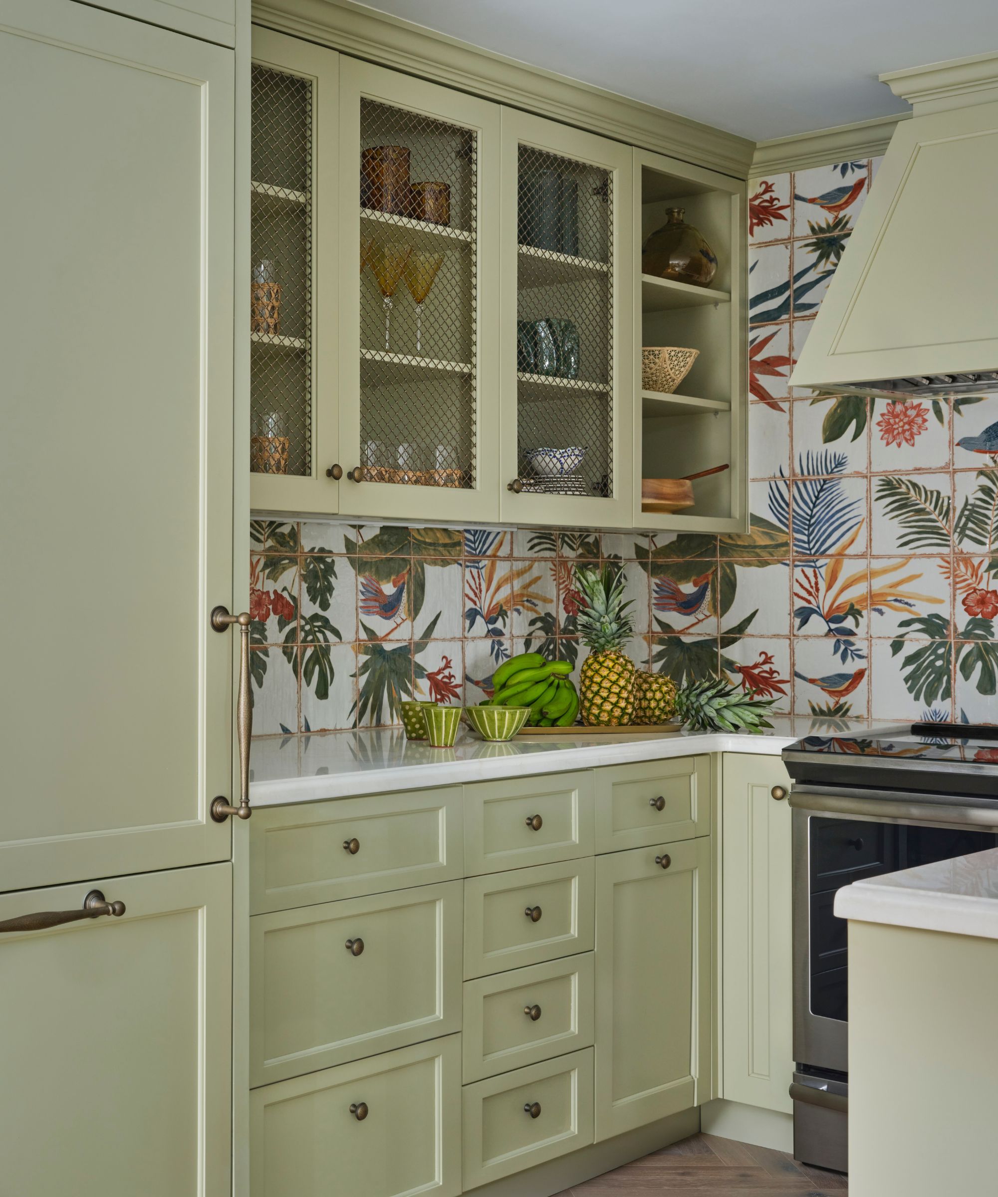 A small kitchen with light green cabinets, tropical print backsplash tiles, and white countertops.
