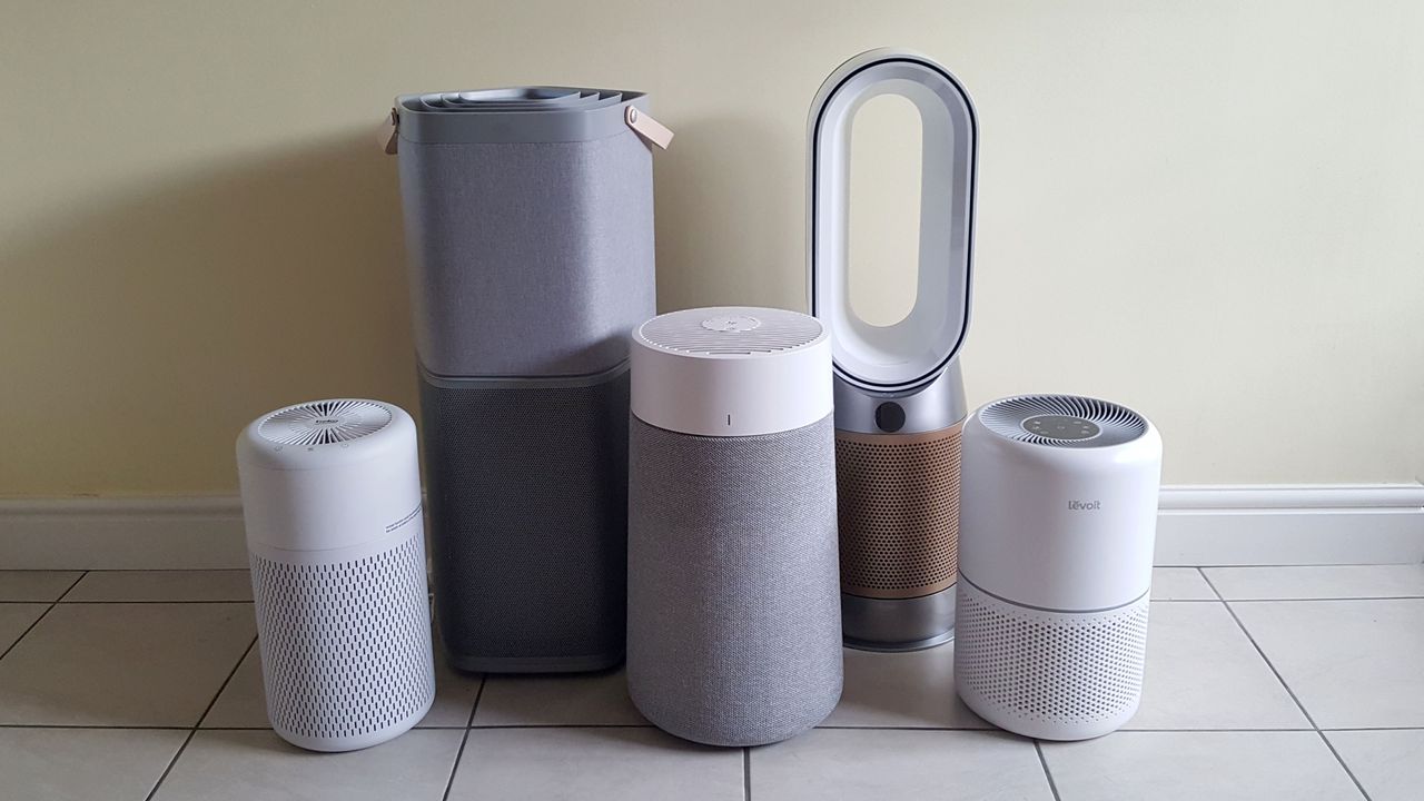 Five air purifiers of various sizes lined up on a tiled floor in a room with a pale yellow wall