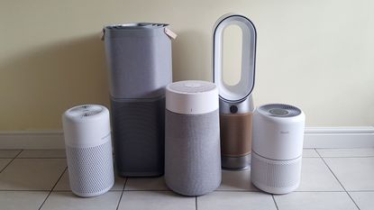 A selection of five air purifiers of various sizes on a wooden table in a room with pale green walls and indoor plants on shelves