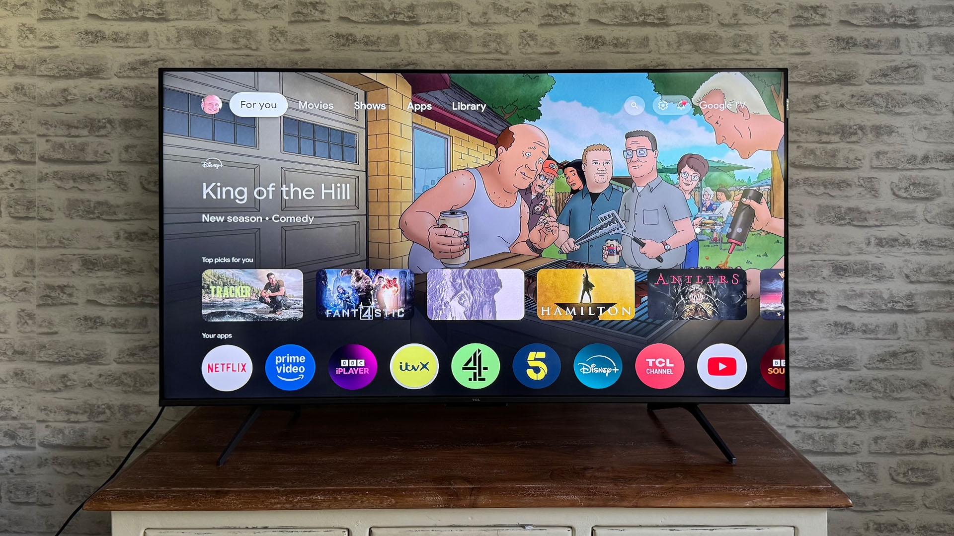 TCL C6KS 50-inch TV in front of brick wall, on screen is Google TV home page and King Of The Hill