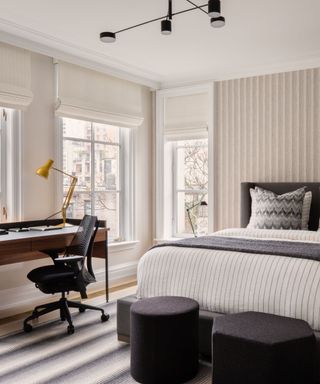 A bedroom with striped neutral wallpaper, two black ottomans, and a wooden desk