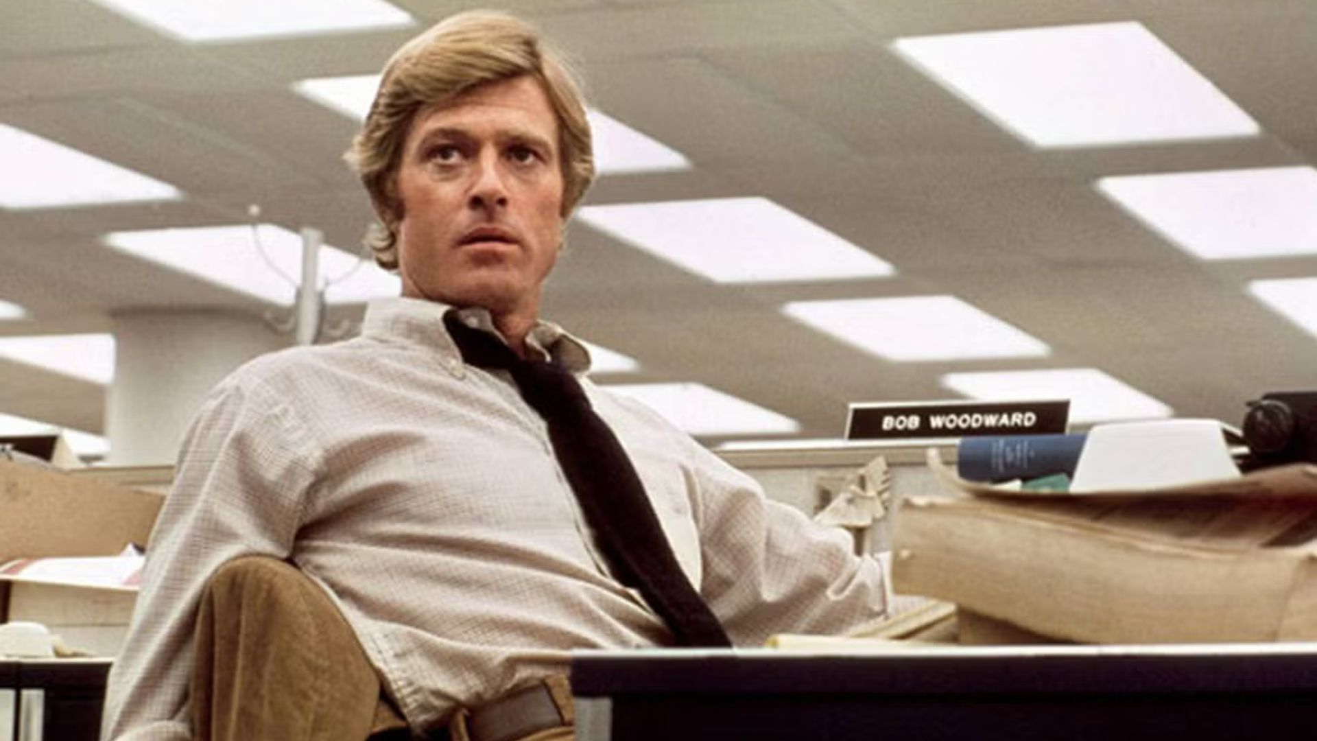 A young Robert Redford sits facing away from a desk