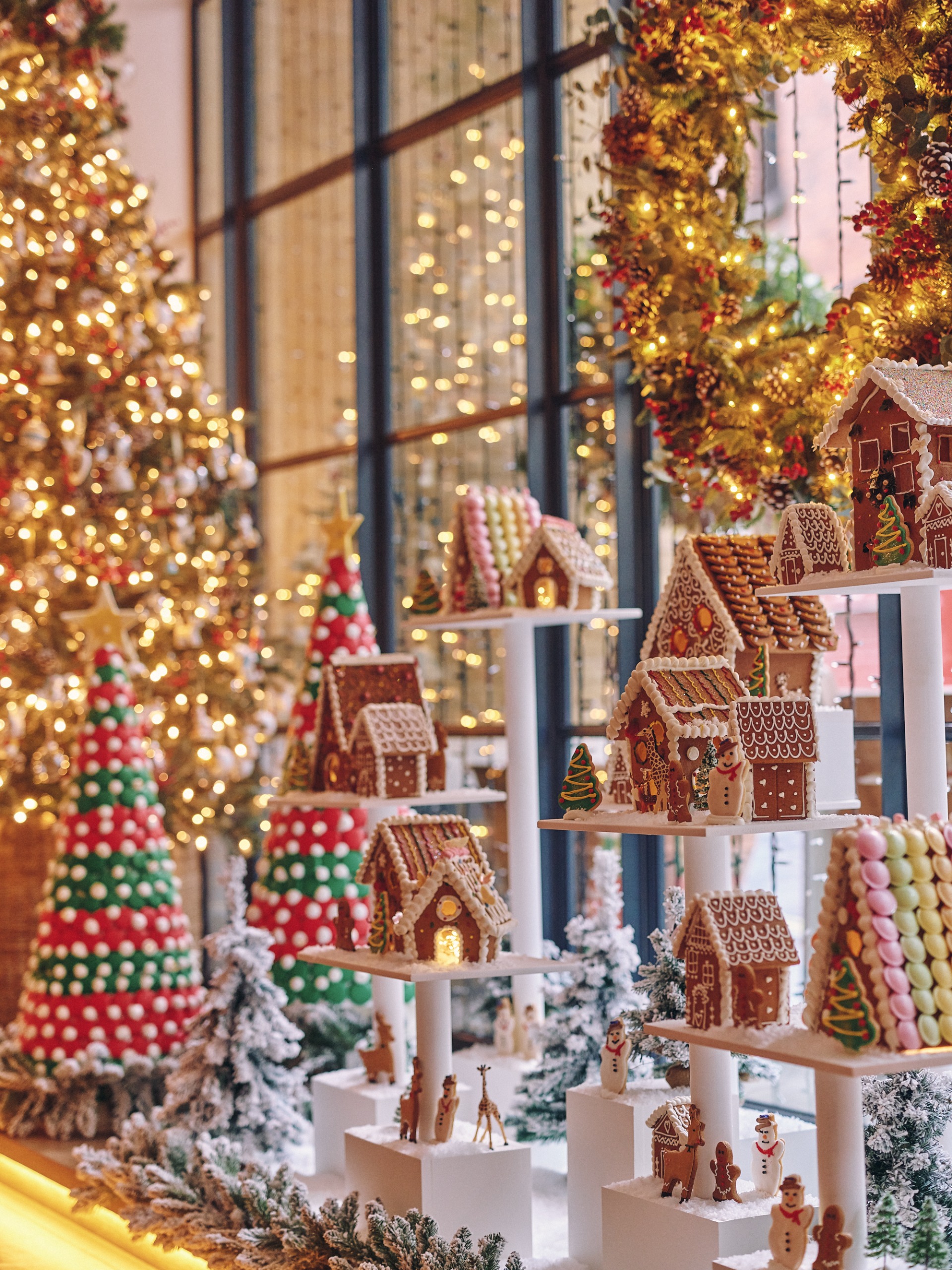 A window display of a gingerbread christmas village on white pedestals at varying heights next to a decorated christmas tree and wreath
