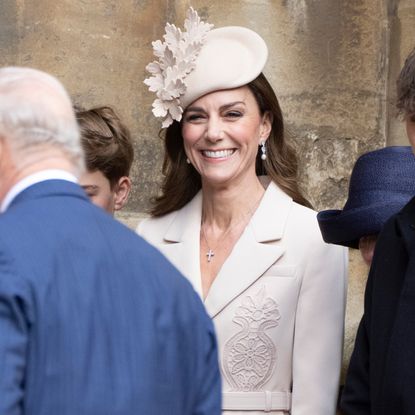 Princess Kate wearing a beige coat and hat and smiling behind The King and Queen