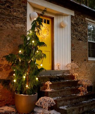 front of a stone cottage decorated for christmas with lights and a christmas tree and a yellow painted front door