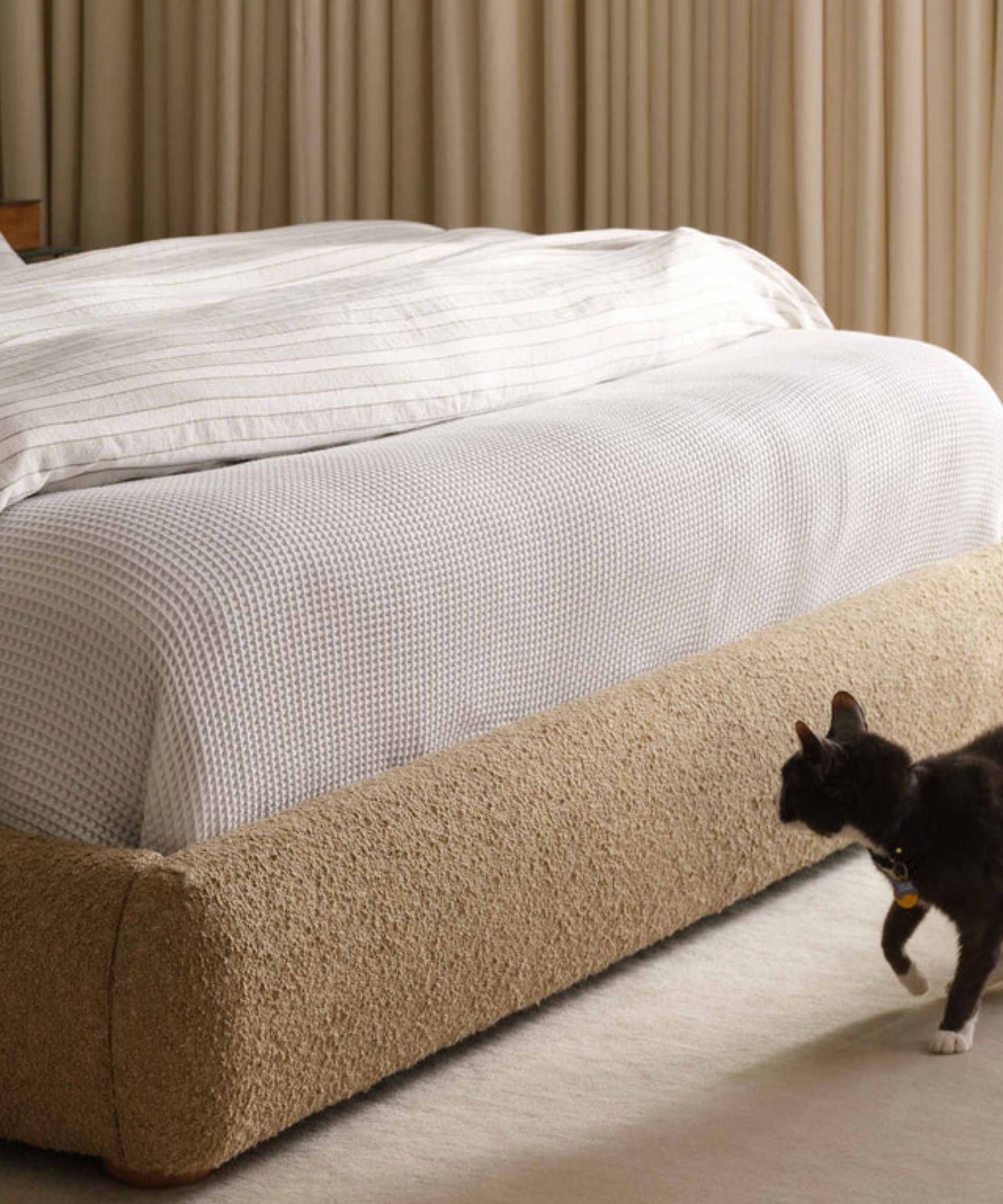 Brooklinen Dreamweave bed blanket tucked into a beige boucle bed frame pictured in a neutral room with beige curtains, white carpet, and a black cat.