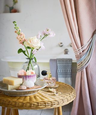 Rattan table in a bathroom with a plate of soaps, vase with flowers and a scent bottle with a bath in the background
