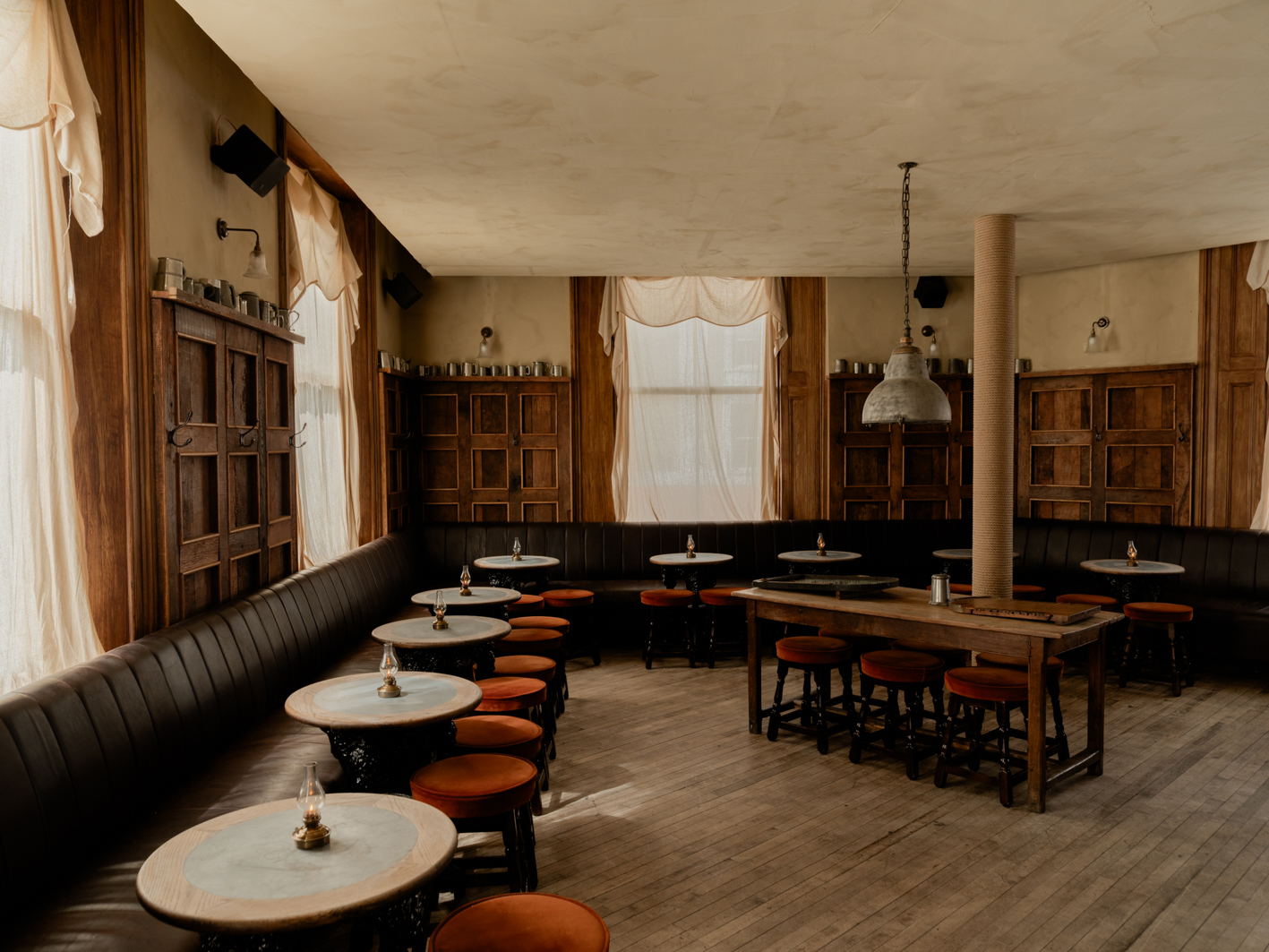 A soulfully decorated pub with velvety round stools, wooden floors, candle-lit atmosphere, and beige curtains.