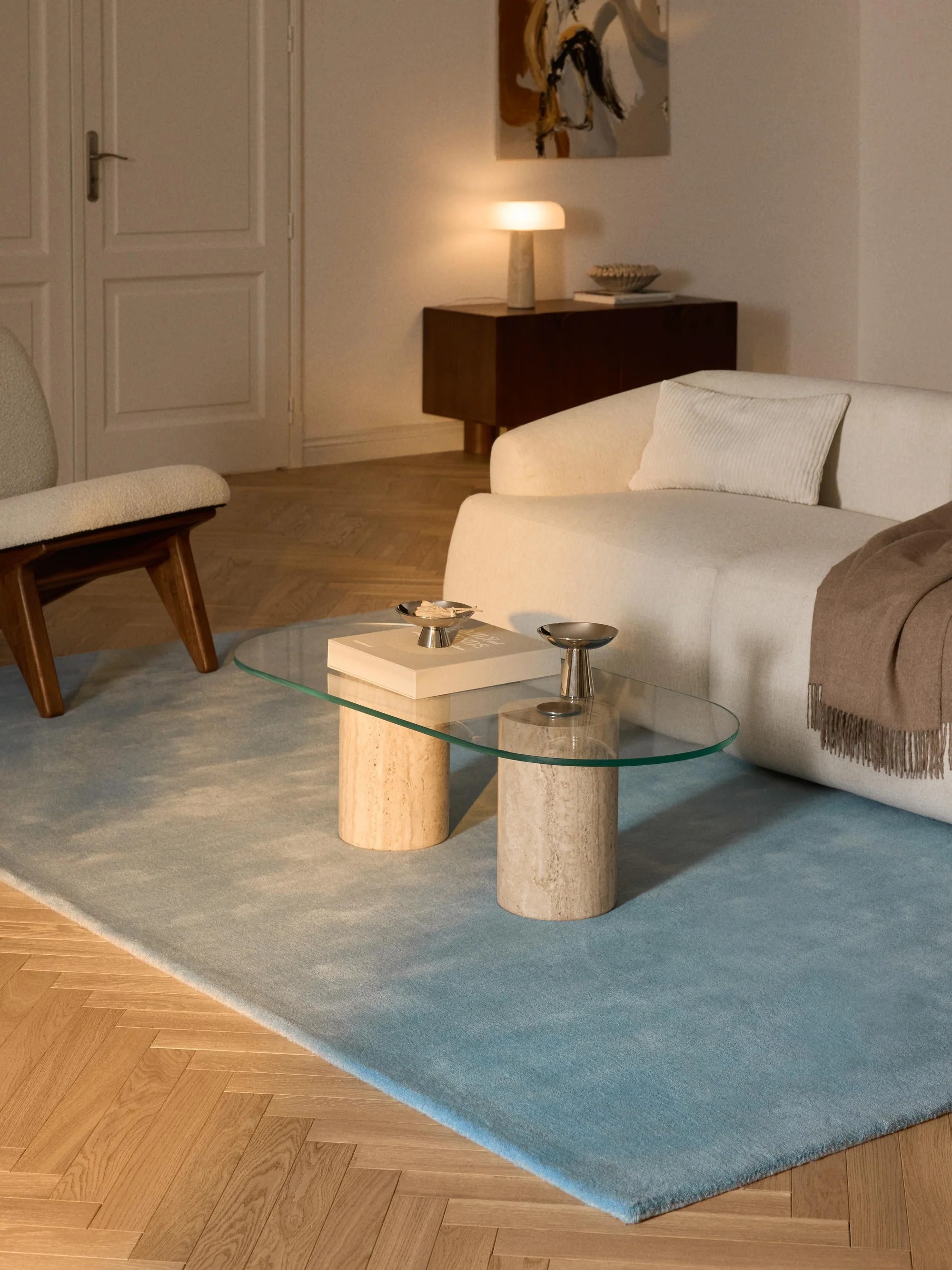 Image of a baby blue rug in a contemporary, luxurious living room. There is a glass and marble coffee table, a white boucle sofa, and a wooden console in the corner with a lamp.