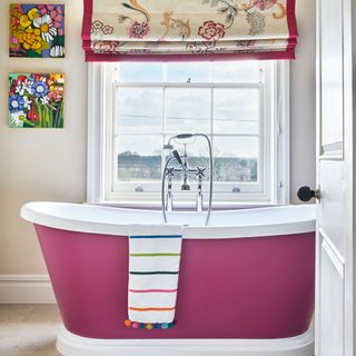 a hot pink bathtub with with edging in front of a window , with a white towel with colourful stripes and bobbles draped over it