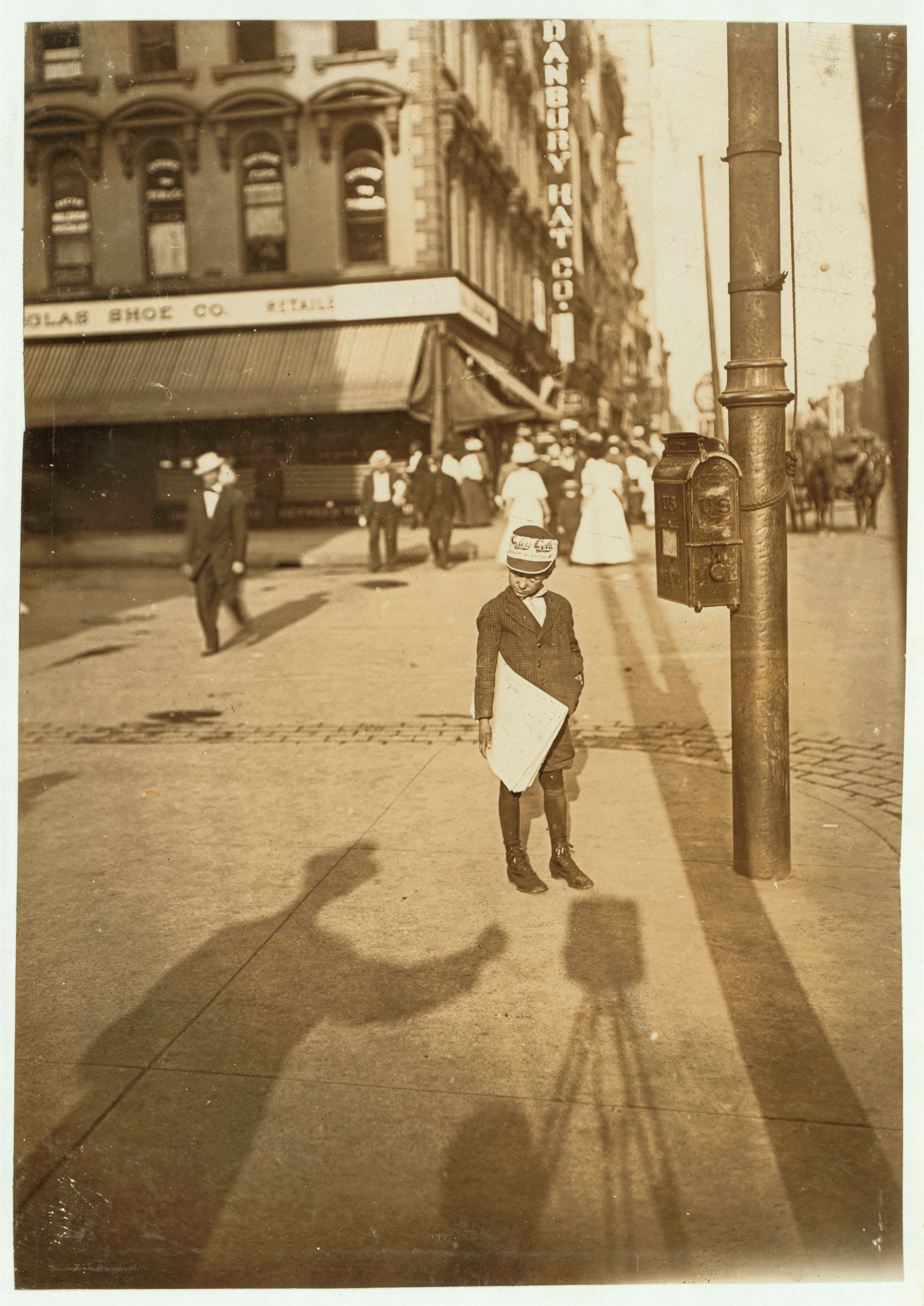 A vintage photograph showing a young newsboy holding papers on a city sidewalk next to a mailbox pole, while the long shadow of the photographer and their tripod stretches out in the foreground.