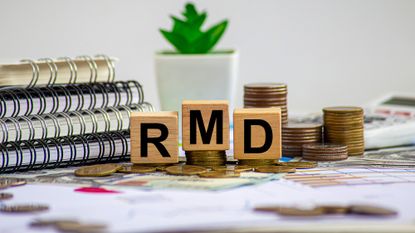 Wooden blocks with the letters RMD spelled out (representing Required Minimum Distribution) sitting on a desk with notebooks and stacked coins