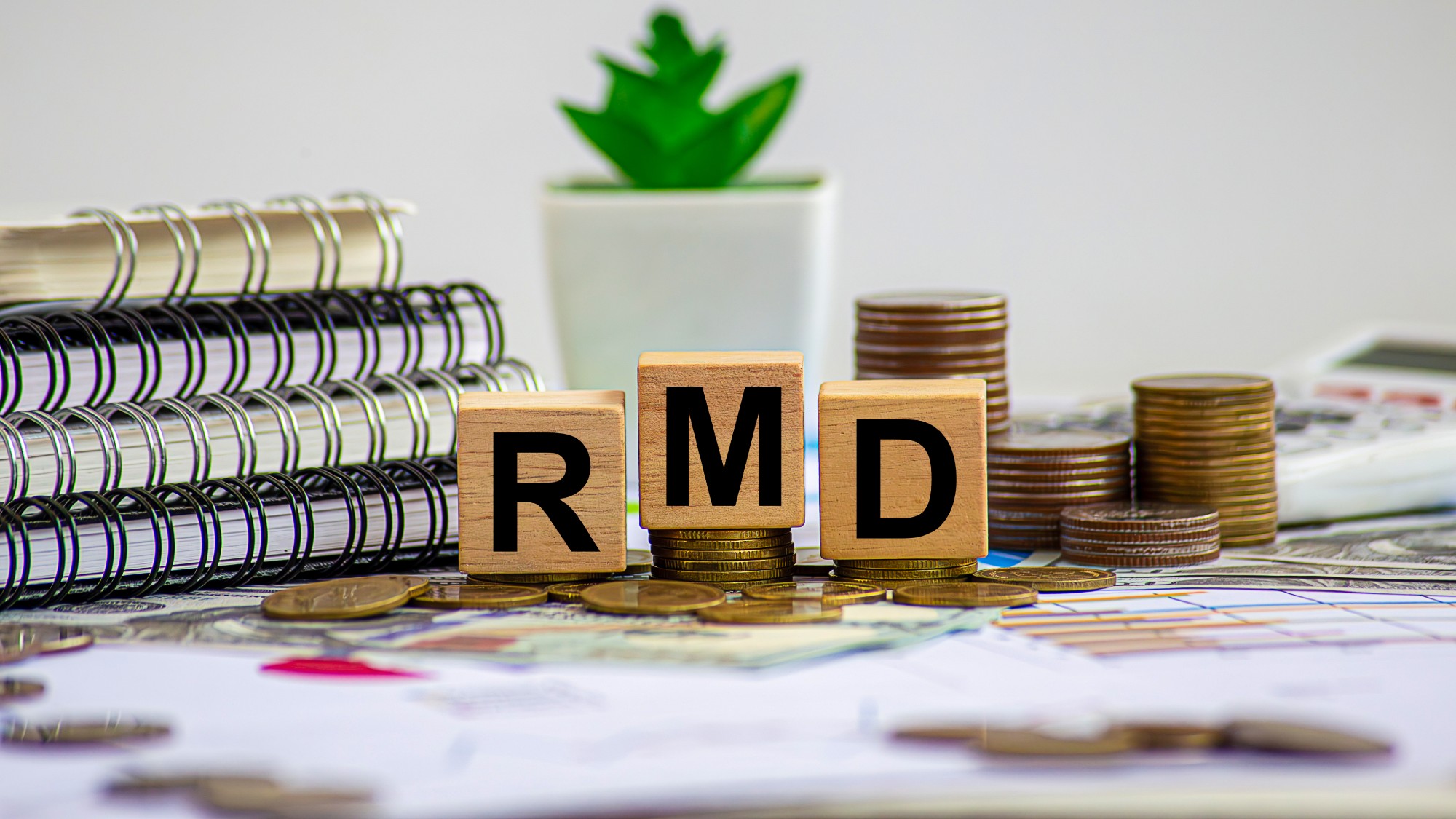 Wooden blocks with the letters RMD spelled out (representing Required Minimum Distribution) sitting on a desk with notebooks and stacked coins