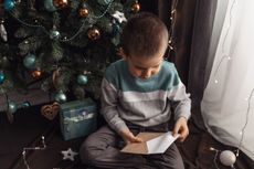 Child opens envelope while sitting under Christmas tree and beside a wrapped gift