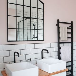 a bathroom with top half of wall in blush pink paint and bottom half with white tiles, with a double sink underneath a mirror with black framing and black chrome taps and towel rail
