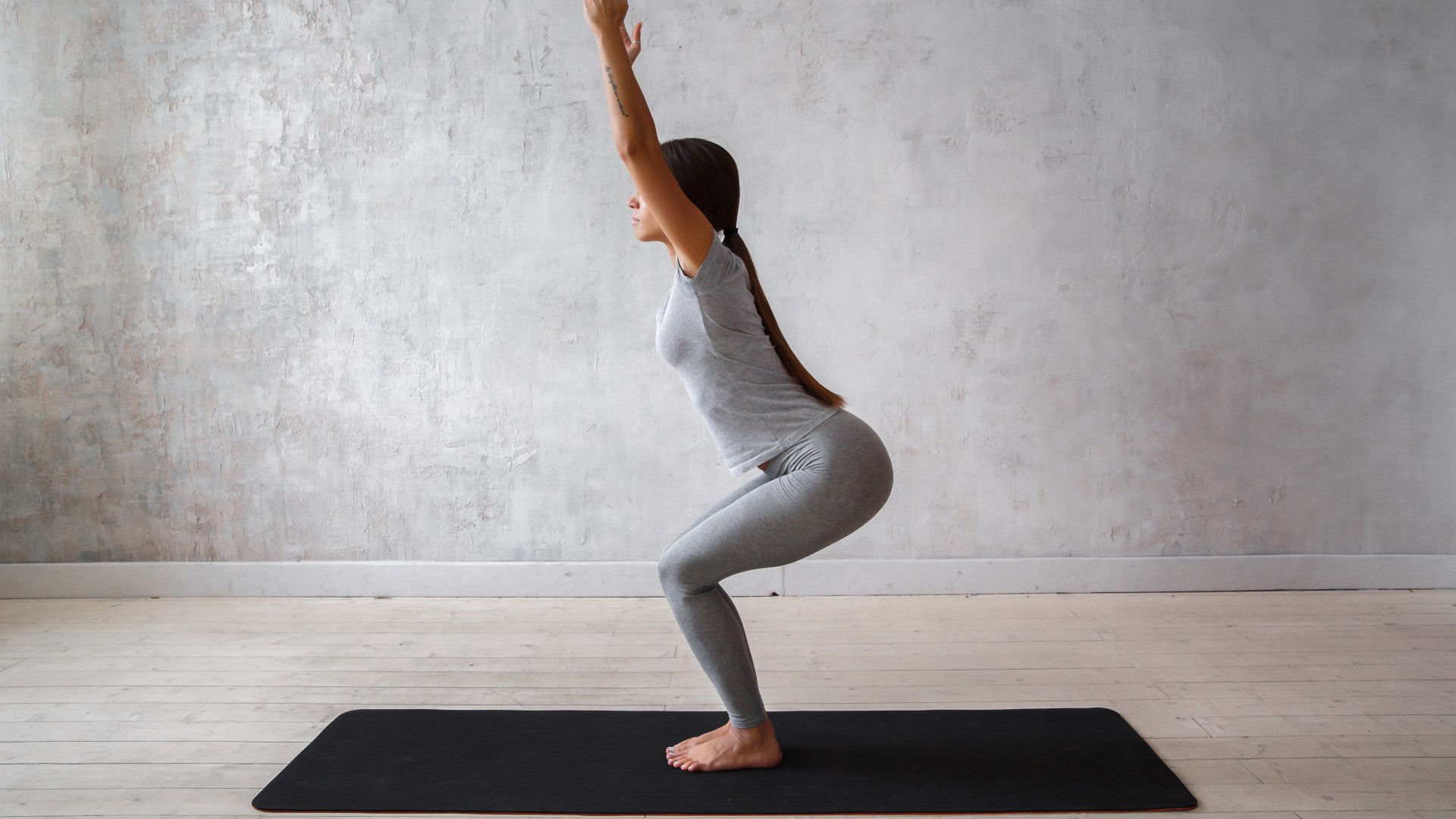 Frau auf ihrer Yogamatte vor grauem Hintergrund mit gebeugten Knien und erhobenen Armen in Stuhlhaltung