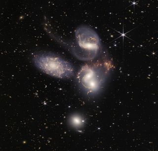 A cluster of three spiral galaxies with hundreds of bright galaxies that look like stars behind them.