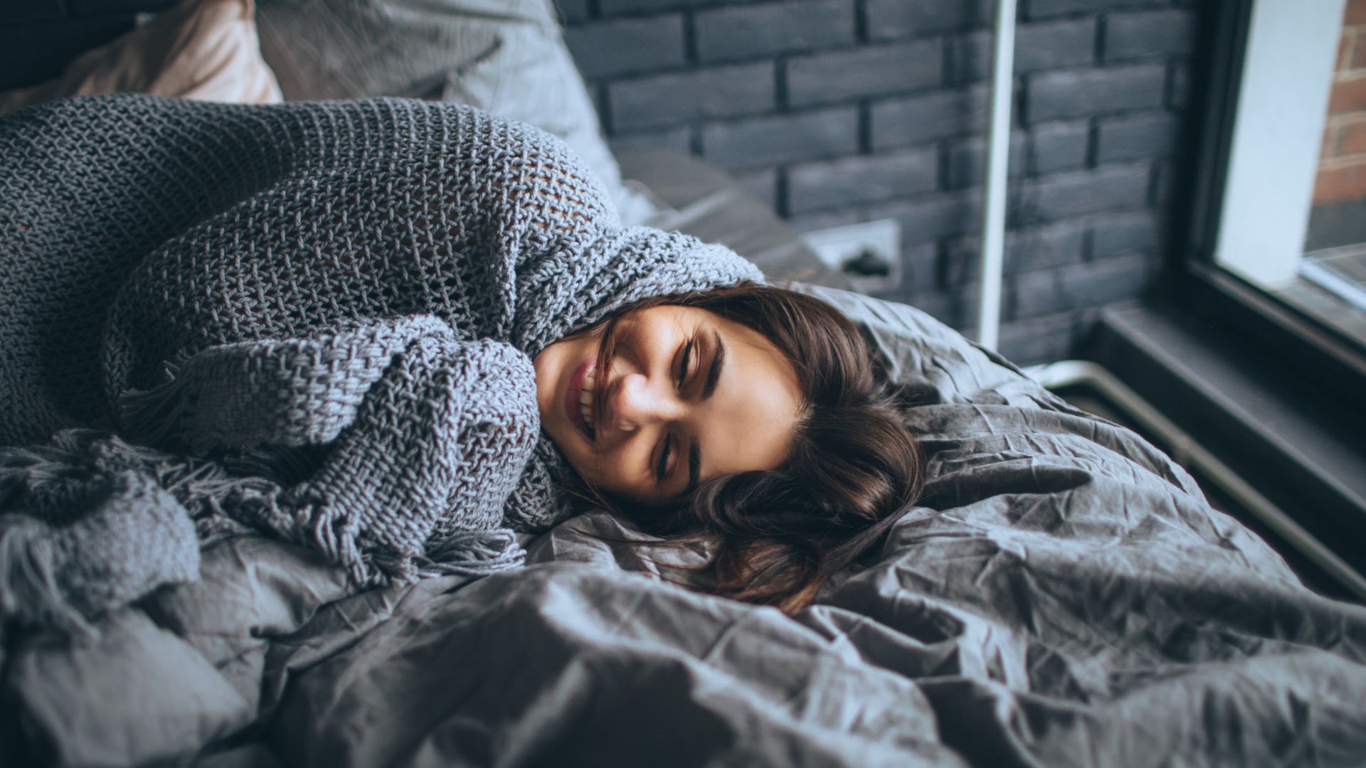 A smiling woman lies in bed wrapped in a gray blanket.