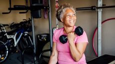 An elderly African American woman sits in a gym, pulling a weight up to the shoulder with one arm.