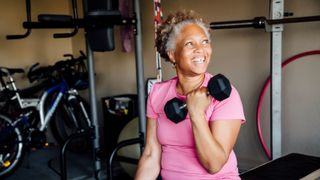 An elderly African American woman sits in a gym, pulling a weight up to the shoulder with one arm.