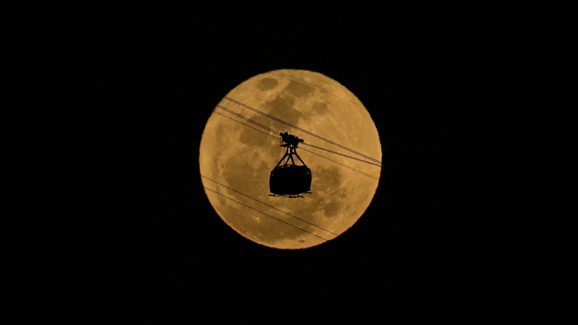 Una luna llena grande y brillante con un ligero tinte naranja en el fondo y la silueta de un teleférico frente a ella.