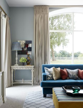 An example of small living room decor ideas showing a blue sofa in a sitting room with blue walls and cream coloured drapes.