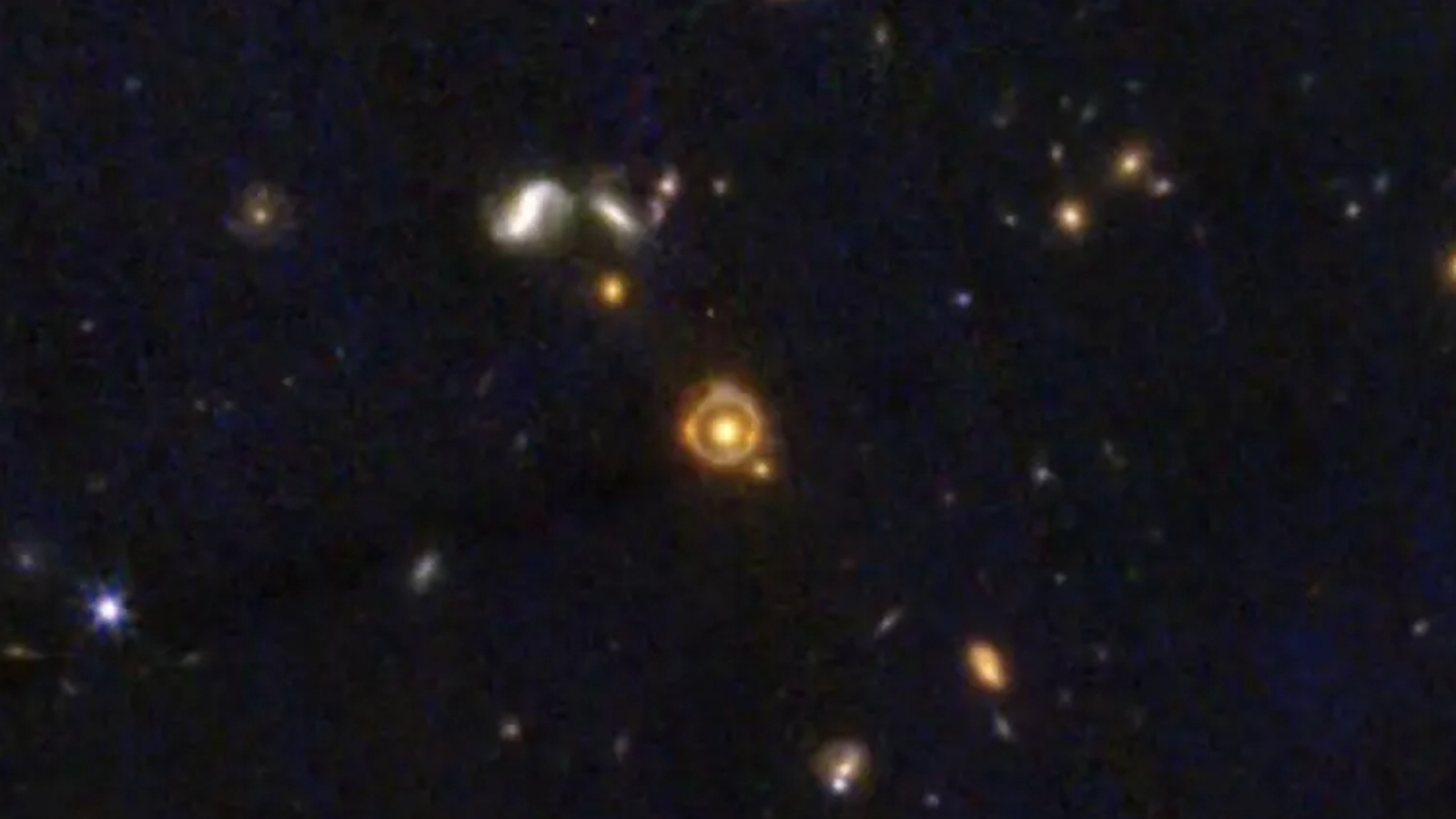Ein Ring aus orangefarbenem Licht umgibt eine gelbe Galaxie in der mit Sternen gefüllten Bildmitte