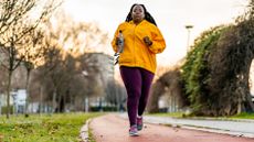 A woman runs on a track holding a water bottle. She wears a bright orange jacket and purple leggings. Behind her we see trees, overgrown hedges and a building.