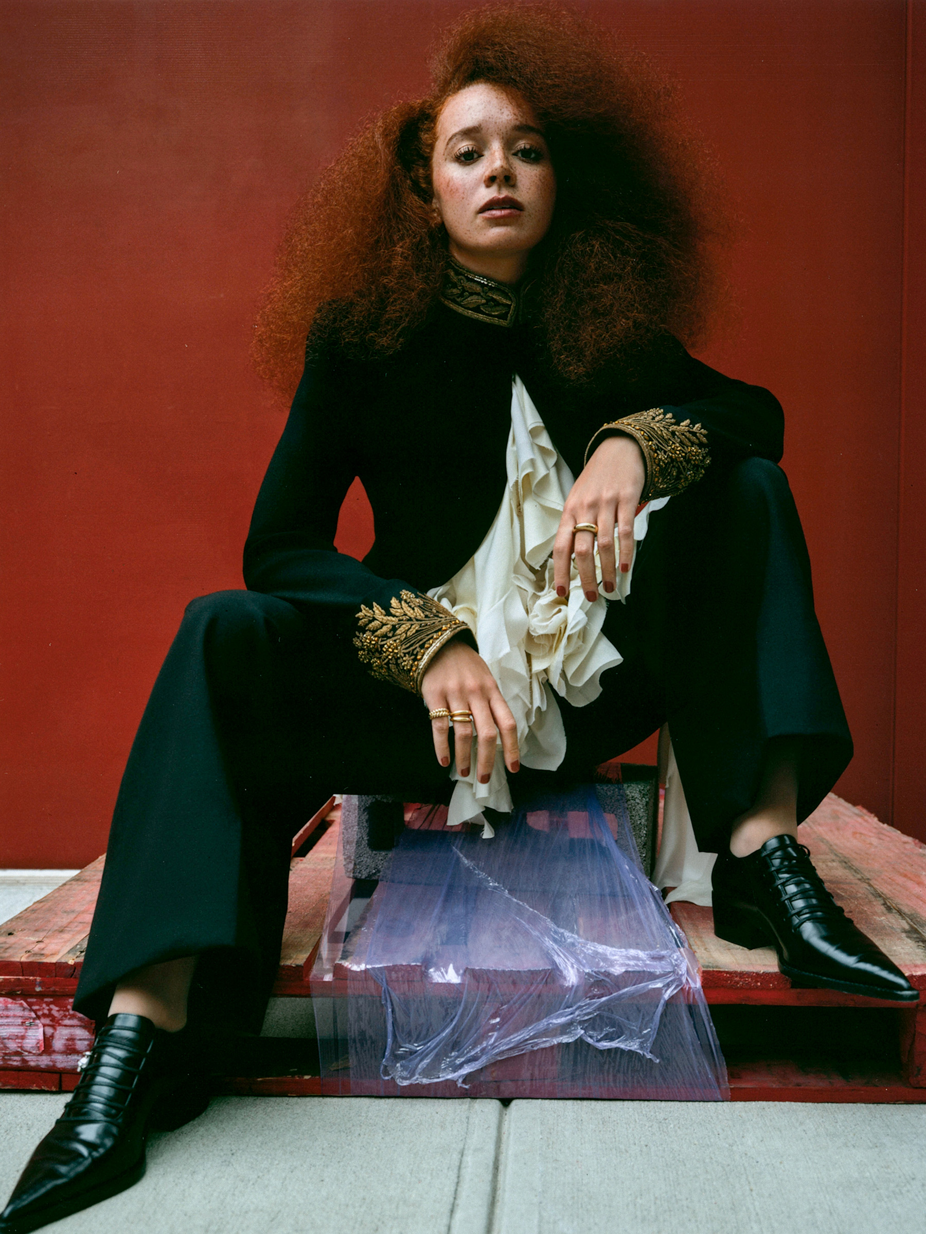 Photo of Erin Kellyman for the Fall Ones to Watch portfolio. She is pictured against a red wall wearing a ruffled Alexander McQueen blouse, a black jacket, black pants, and shoes with Meuchner jewelry.