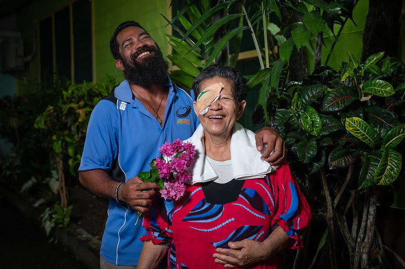 A man with a full beard and blue polo shirt stands next to a smiling woman with an eye patch, red patterned dress, and white towel draped around her neck, holding a small bouquet of pink flowers.