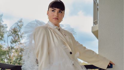 Devery Jacobs stands on a balcony in a cream suit with white tulle all over