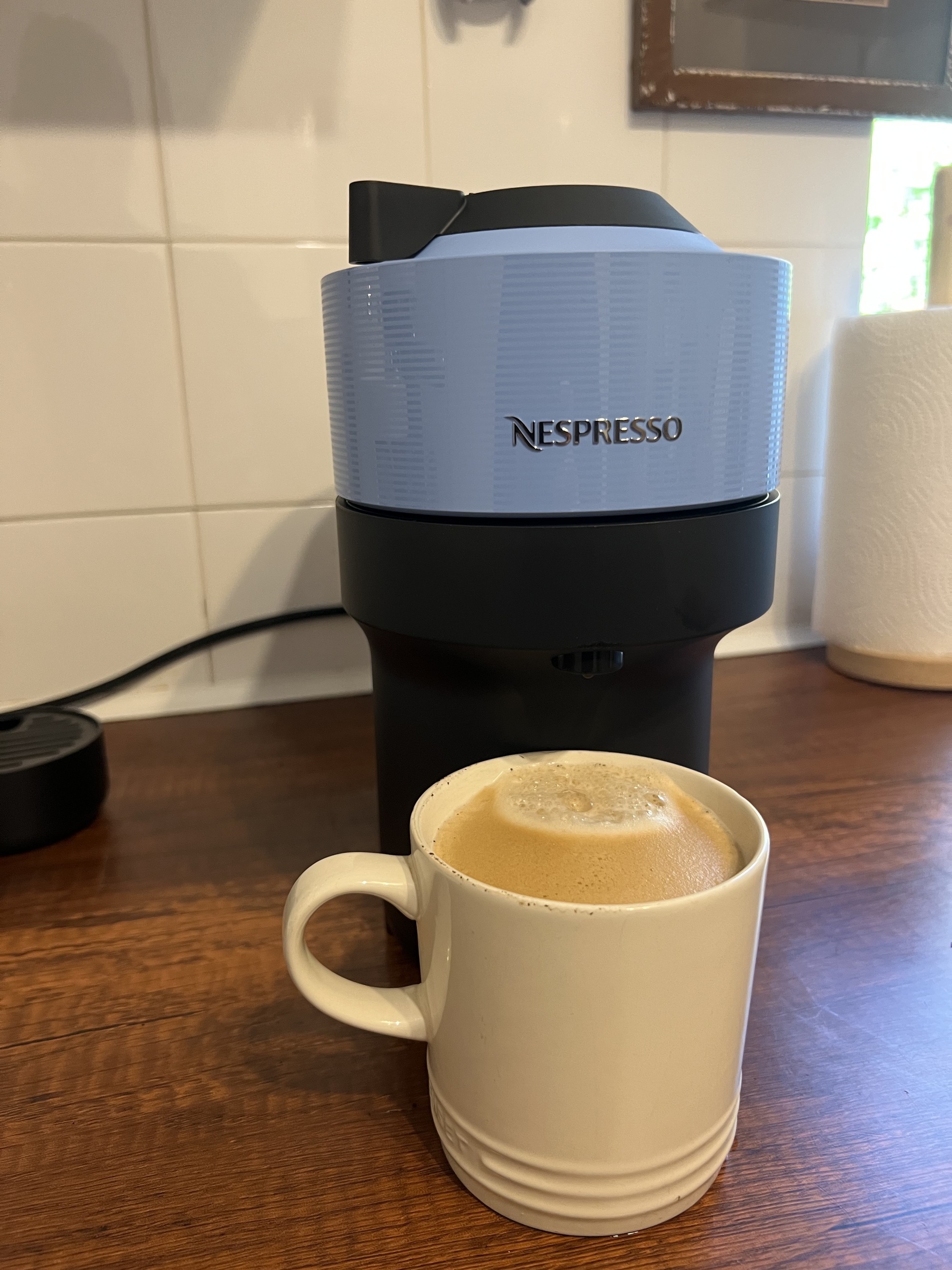 Image of a white coffee mug full of coffee in front of a blue and black Nespresso Vertuo Pop machine.
