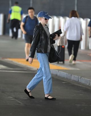 Julia Garner am LAX trägt eine blaue Baseballkappe, ein gestreiftes T-Shirt, eine Lederjacke, Jeans und Mary-Jane-Flats.