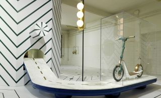 View of a green, white and bronze coloured electric scooter in a clear display box. The scooter sits in a space with patterned flooring, sphere lighting that resembles a traffic light and a patterned structure with an elevated path that resembles a road