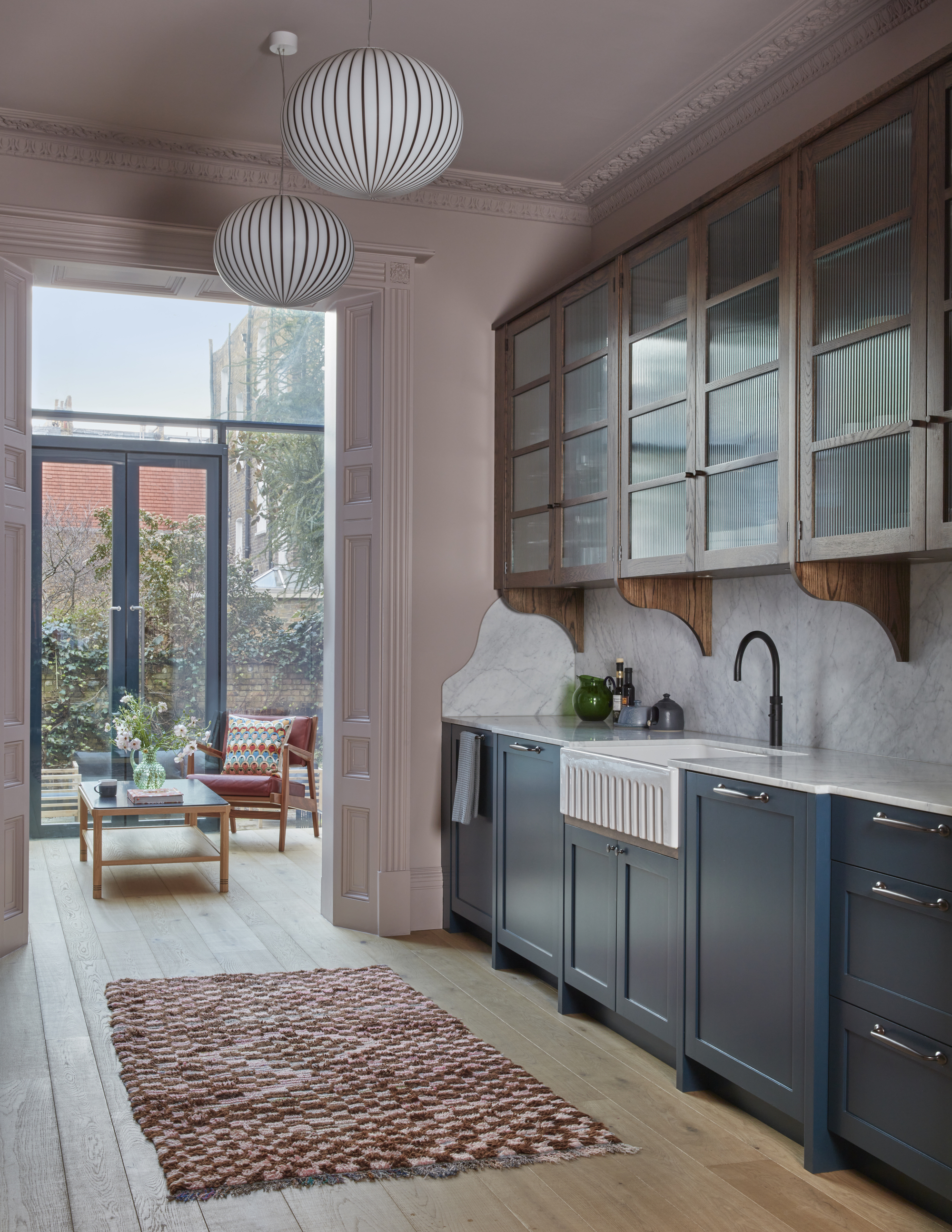 navy blue galley kitchen with pink walls and ceiling, timber floors, and check floor rug, and round pendant lights and a hallway leading through to a sunroom with an armchair and coffee table