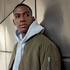 Male model leaning against a wall wearing a light grey hoodie and khaki bomber jacket sold at Burton