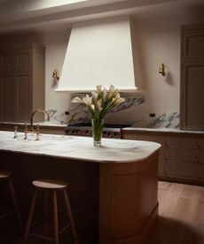 Neutral kitchen with oval island with wooden base and marble top and wooden stools. Large white range good, painted green cabinetry, marble backsplash