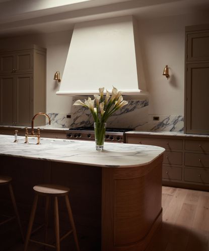 Neutral kitchen with oval island with wooden base and marble top and wooden stools. Large white range good, painted green cabinetry, marble backsplash