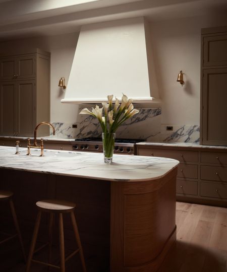 Neutral kitchen with oval island with wooden base and marble top and wooden stools. Large white range good, painted green cabinetry, marble backsplash